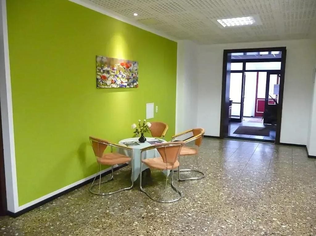 Una habitación con pared verde, un cuadro, una mesa con sillas y una puerta que da a otra zona.