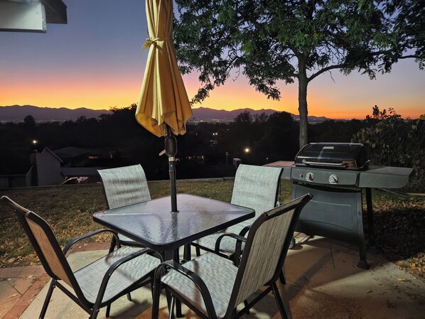 An outdoor patio with a table and chairs, a grill, and a view of the sunset over mountains.