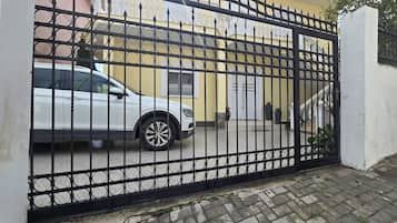 Een omheind toegangspunt met een witte auto erin, een wit gebouw met een balkon, en een geplaveid pad dat naar de ingang leidt.