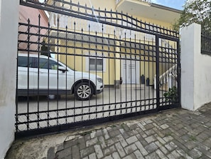 A gated entrance with a white car parked inside, a white building with a balcony, and a paved pathway leading to the entrance.