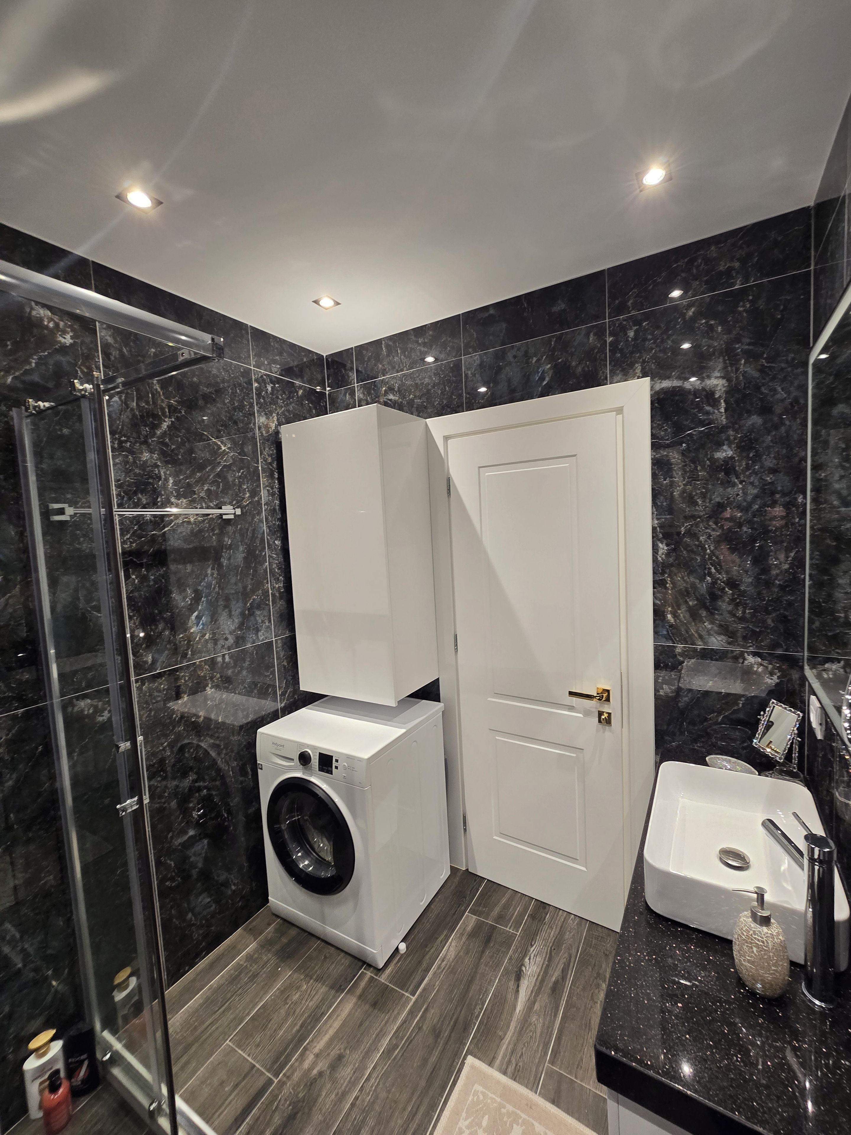 Une salle de bain moderne avec une douche à parois de verre, un lave-linge encastré, une porte blanche et un lavabo doté d’un plan de travail en marbre.