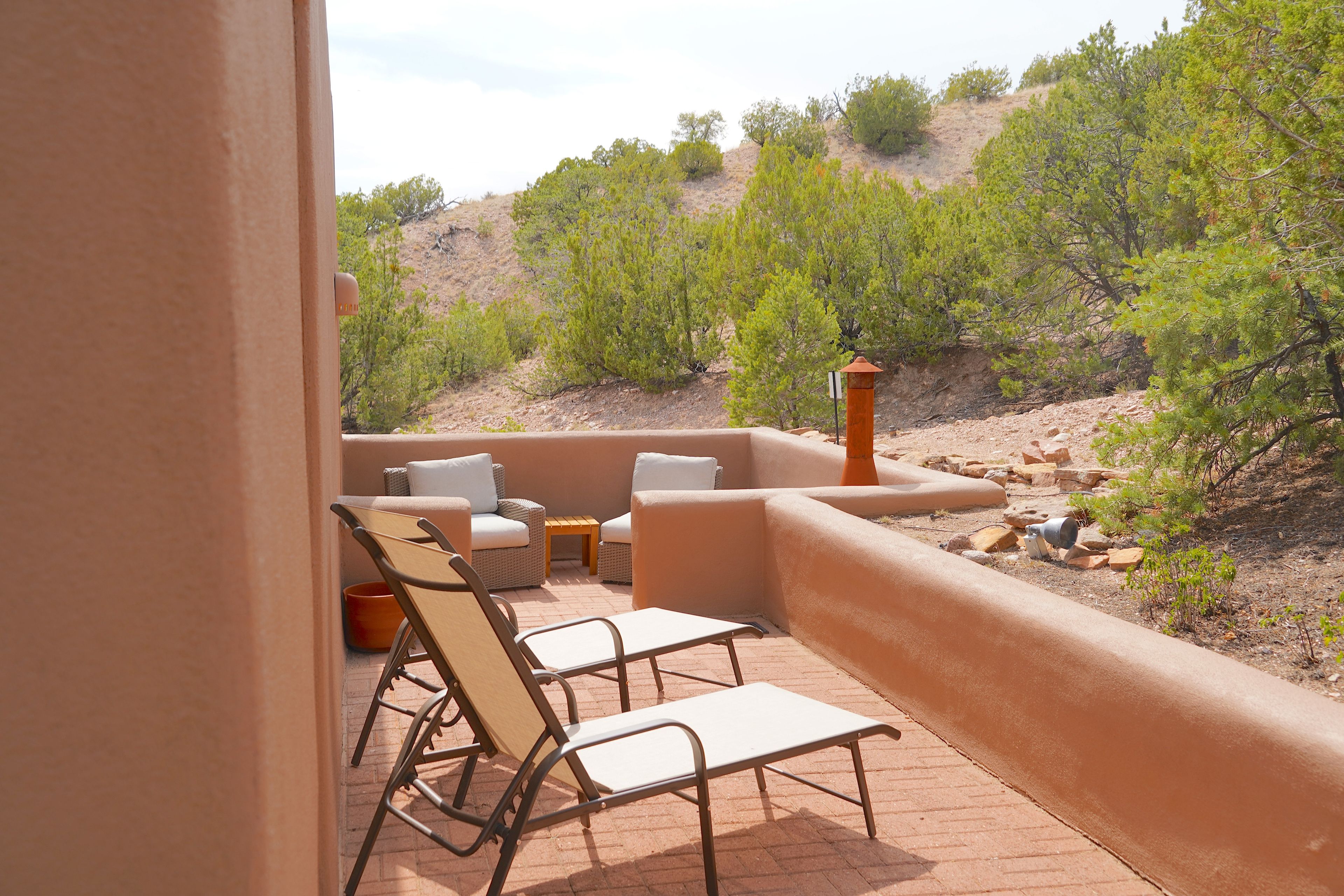 An outdoor patio with a seating area, including a table and chairs, overlooking a natural landscape with trees and a hill.