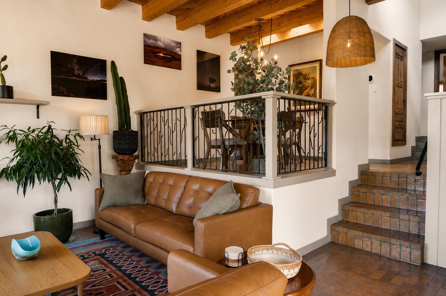 A living room with a brown leather sofa, a coffee table, and a potted plant. There are framed pictures on the wall and a staircase leading to another level.
