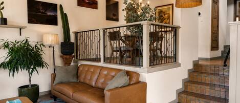 A living room with a brown leather sofa, a coffee table, and a potted plant. There are framed pictures on the wall and a staircase leading to another level.