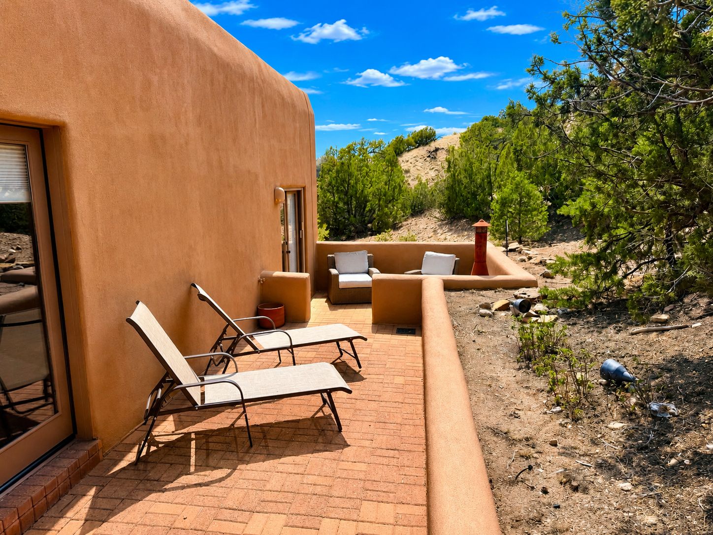 A patio with lounge chairs and a small table, a door, and a view of a natural landscape with trees and hills.
