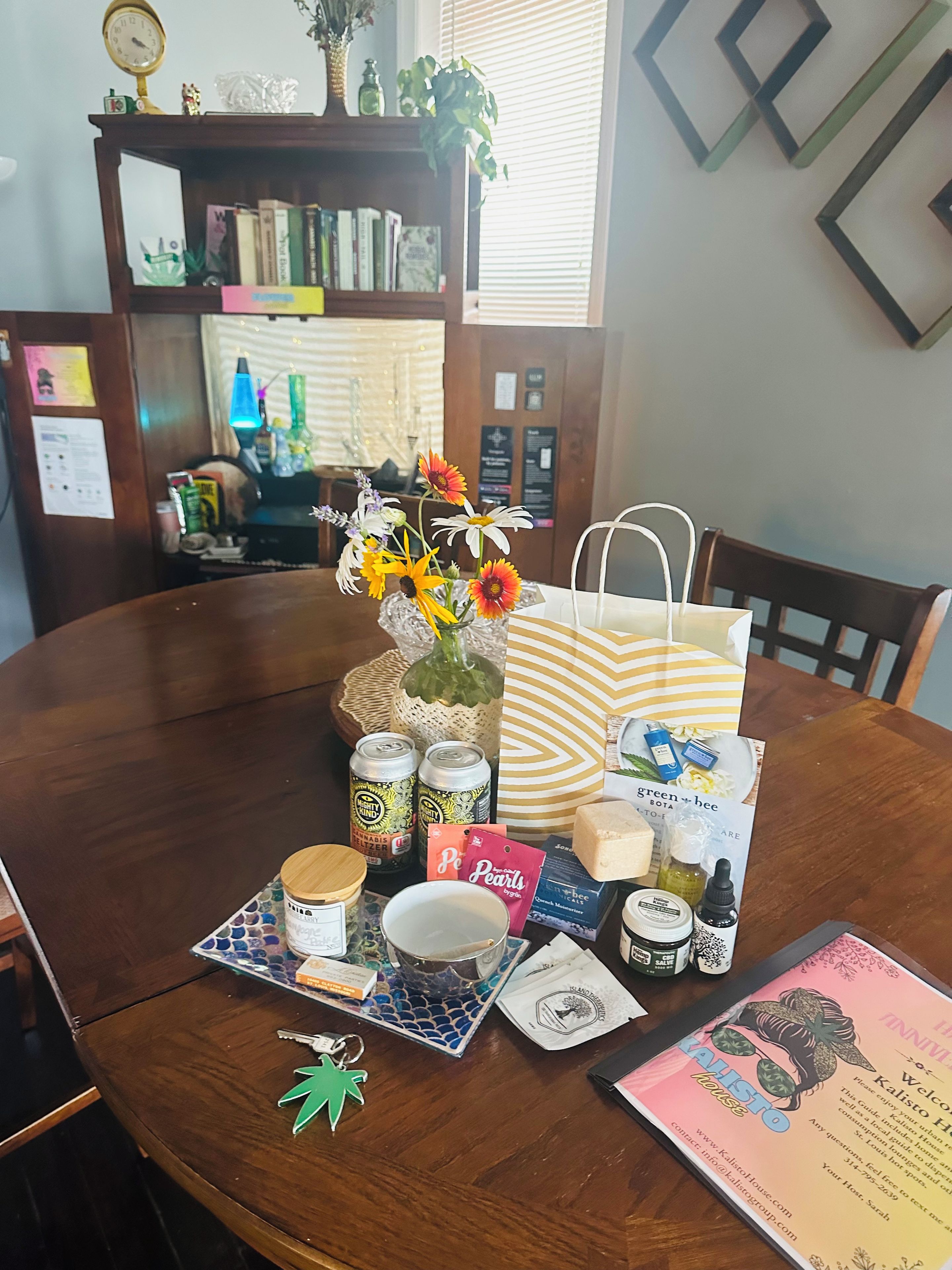 A wooden dining table with a vase of flowers, a striped bag, and various items including cans, a jar, and a keychain.