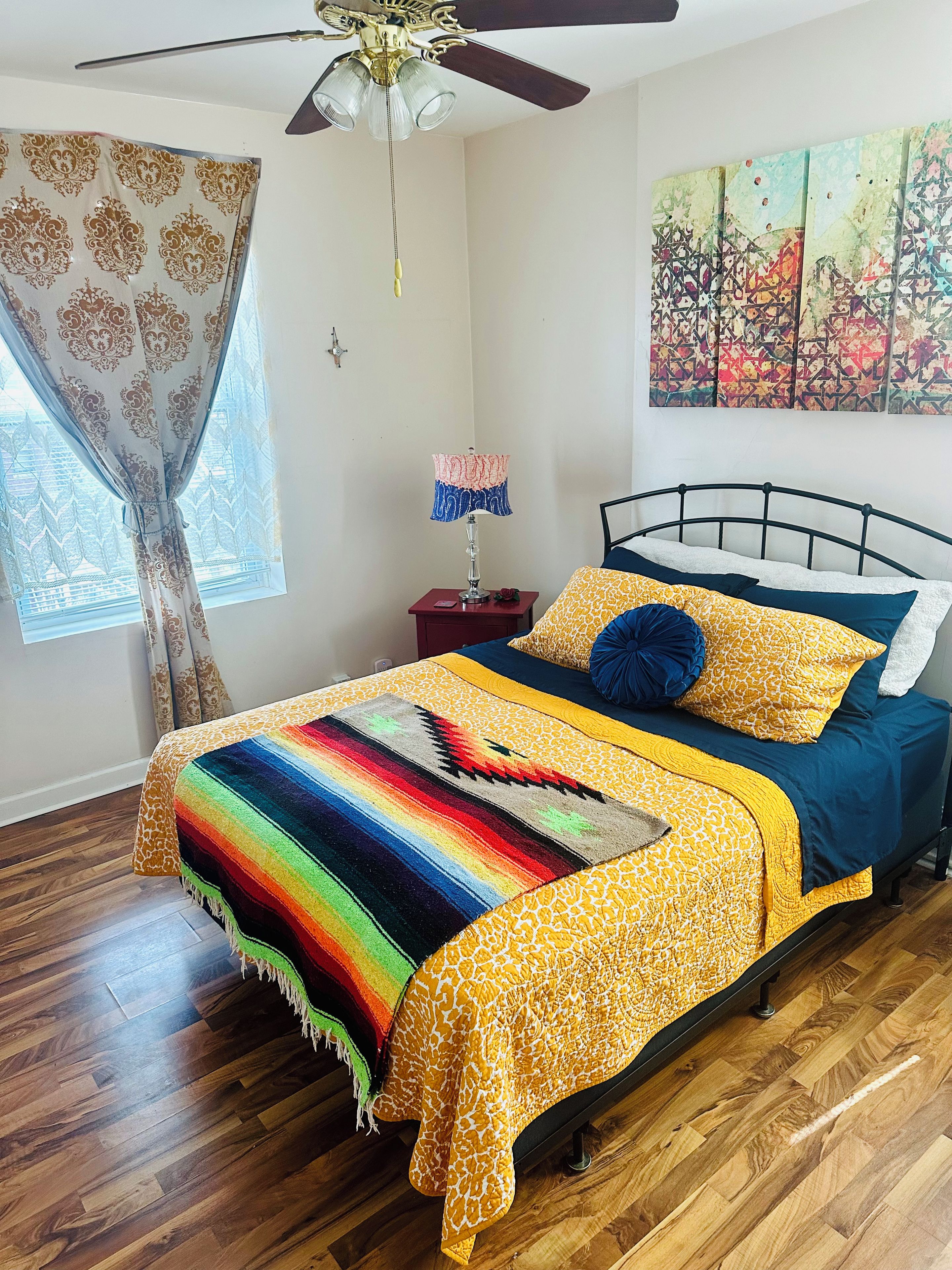 A bedroom with a bed, a nightstand, a lamp, a ceiling fan, a window with curtains, and a wall painting.