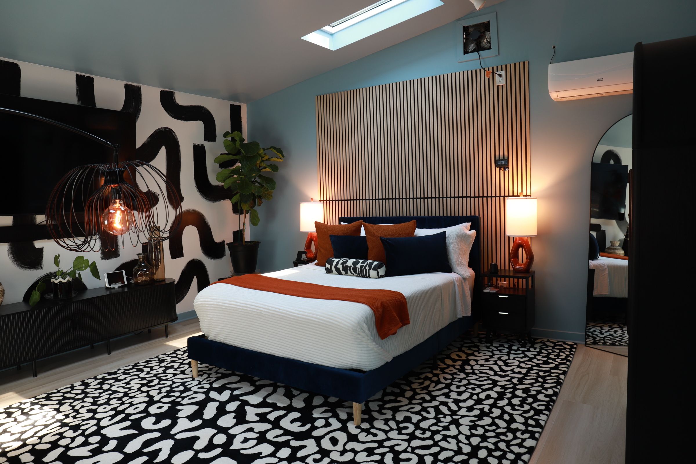 A modern bedroom with a skylight, a bed with multiple pillows, a patterned rug, and a wall with vertical lines.