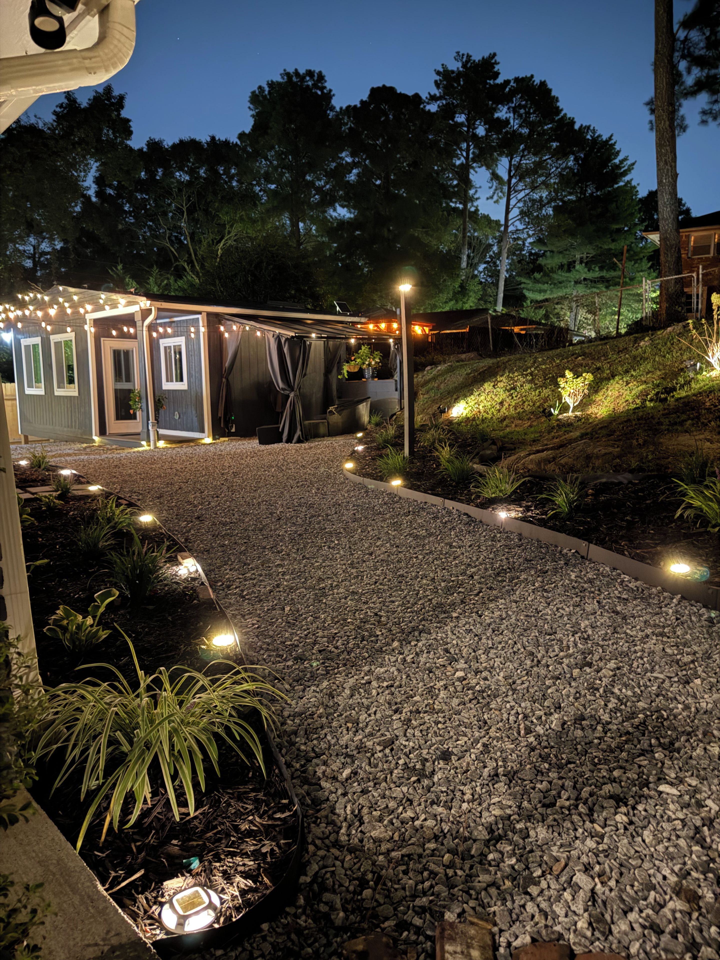 A gravel pathway leading to a building with string lights, surrounded by plants and trees.