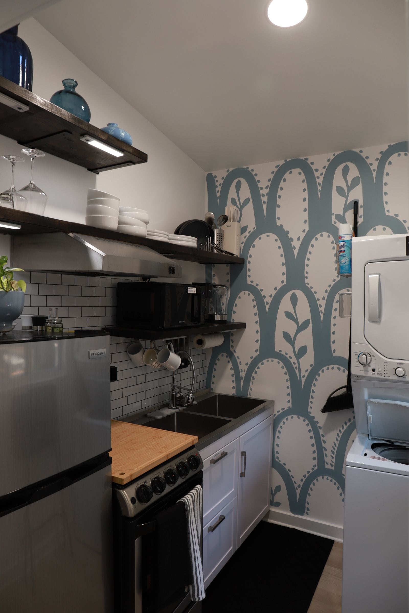 Une cuisine équipée d’une cuisinière, d’un four, d’un four à micro-ondes et d’un réfrigérateur. Des étagères situées au-dessus de la cuisinière servent à ranger la vaisselle, et un vase bleu y est également posé.