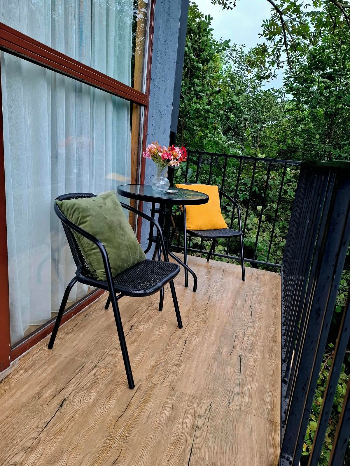 Uma varanda com piso de madeira, uma cadeira de metal preto com almofada verde, uma almofada amarela e uma pequena mesa redonda com um vaso de flores.