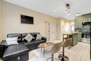 A modern living room with a black leather sofa, a small wooden coffee table, and two bar stools. - Manzil Suite | Cozy 2BR Private Retreat (Maple Ridge)