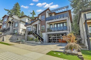 A modern multi-story residential building with a garage entrance, outdoor stairs, and a landscaped area with a small tree and decorative pots. - Manzil Suite | Cozy 2BR Private Retreat (Maple Ridge)