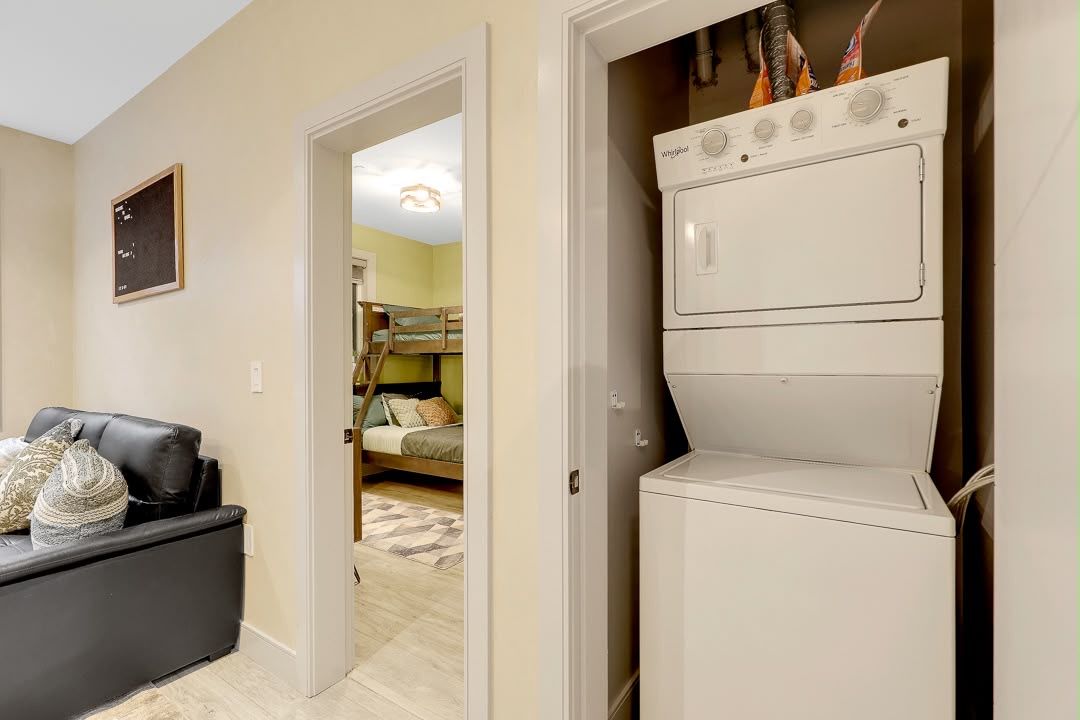 A laundry room with a stacked washer and dryer, a couch with pillows, and a door leading to a bedroom with a bunk bed.