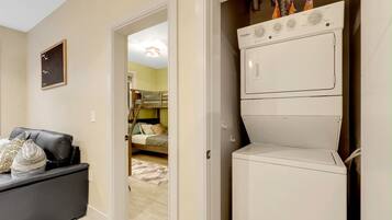 A laundry room with a stacked washer and dryer, a couch with pillows, and a door leading to a bedroom with a bunk bed.
