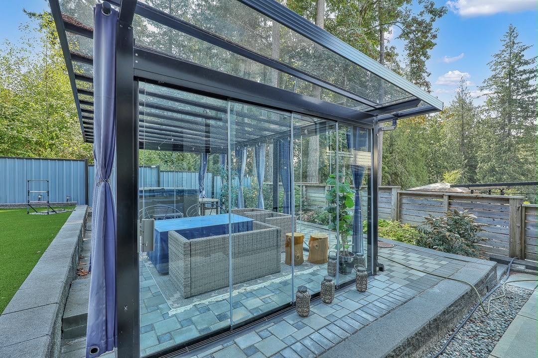 A modern glass-enclosed patio with outdoor furniture, a hot tub, and a wooden deck.
