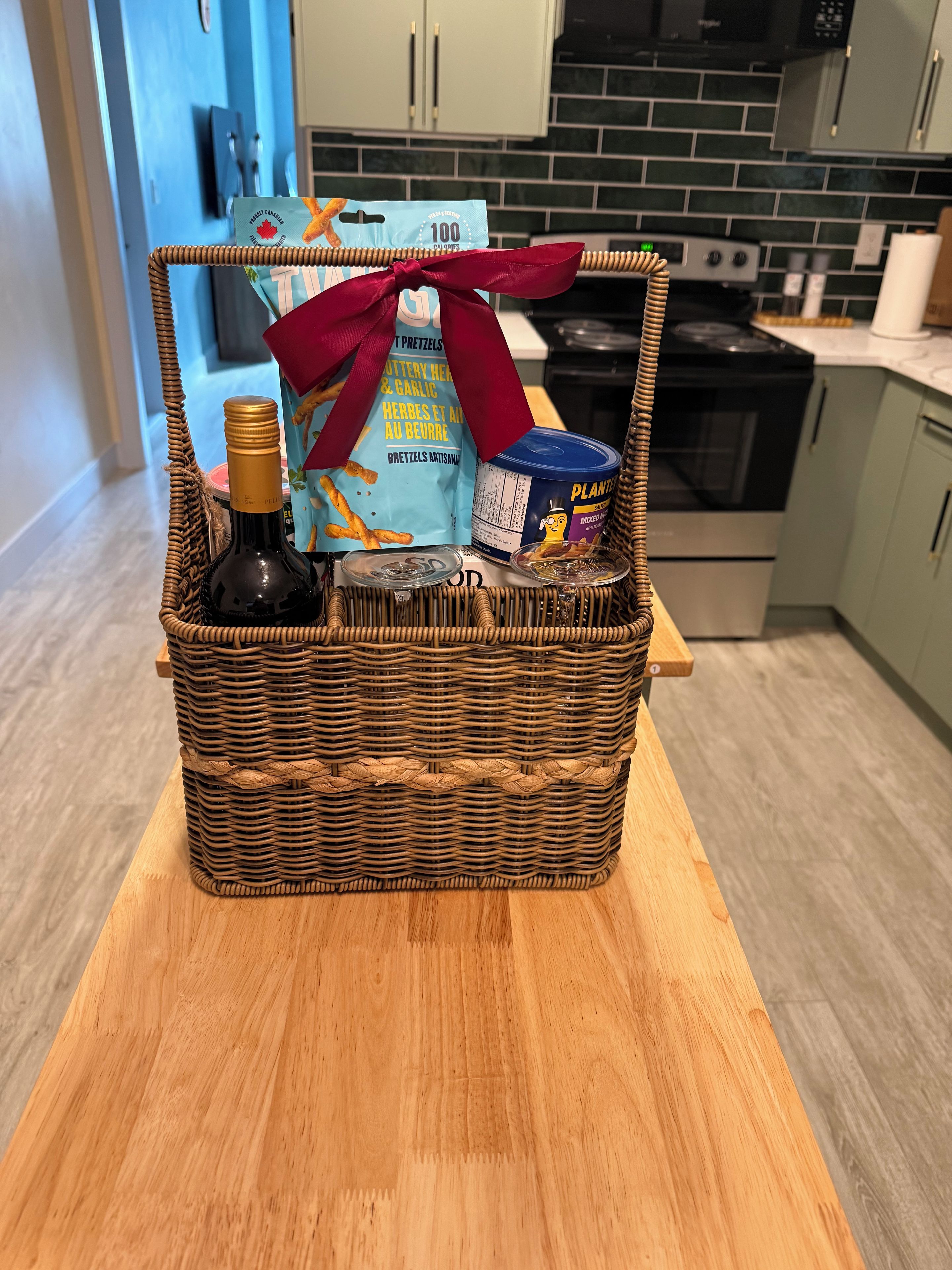 A wicker basket on a wooden table containing a bottle, a bag of pretzels, a can of Plantronics, and a red ribbon.
