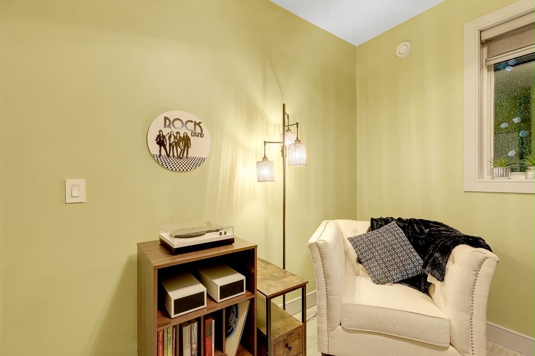 A room with a beige armchair, a small wooden shelf with books, a wall-mounted light fixture, and a framed picture on the wall.