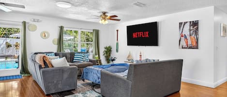 A modern living room with a flat-screen TV, a ceiling fan, and a large window with curtains.