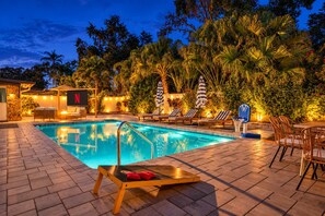 A swimming pool with lounge chairs and umbrellas, surrounded by tropical plants and a stone wall. - Heated Pool + Game Room, Near Clearwater Beach (Largo)