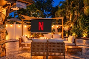An outdoor seating area with wicker furniture, a fire feature, and a large screen displaying the Netflix logo. - Heated Pool + Game Room, Near Clearwater Beach (Largo)