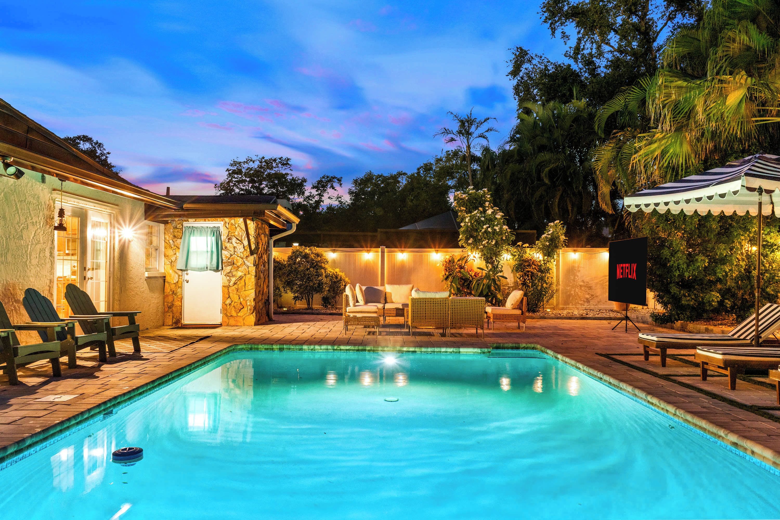 A swimming pool with lounge chairs and a seating area, surrounded by trees and a building with stone walls.
