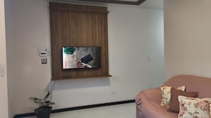 Una sala de estar con un televisor montado, un mueble de madera, una planta en maceta y un sofá con cojines estampados.
