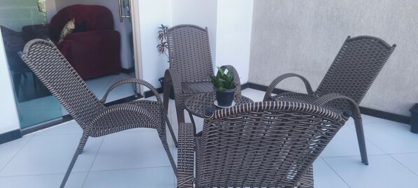 Un área de estar exterior con muebles de mimbre, cuatro sillas y una mesa, una planta en maceta y una silla roja al fondo.
