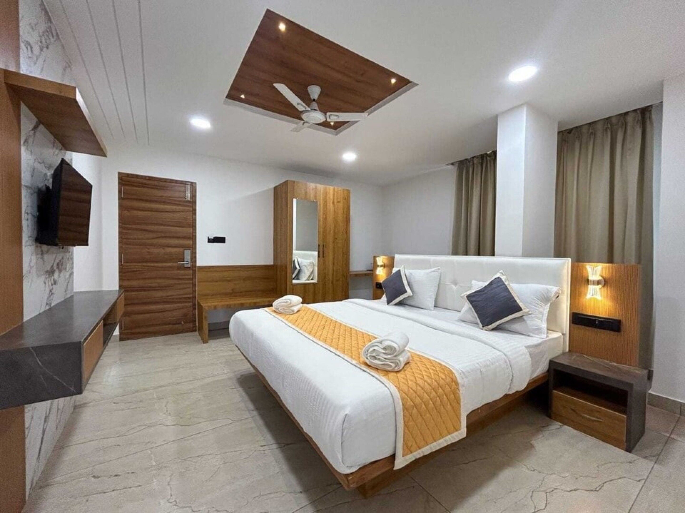 A hotel room with a bed, a bedside table, a mirror, a ceiling fan, and a wall-mounted TV.