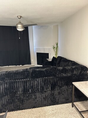 Un salon avec un canapé d’angle noir, une cheminée, un ventilateur de plafond et une petite table.