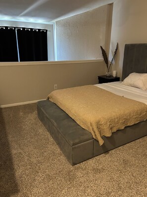Une chambre à coucher avec un lit, une table de chevet, une fenêtre avec des rideaux et un sol recouvert d’une moquette.