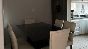 Un espace repas moderne avec une table à plateau en verre et quatre chaises, une bibliothèque en bois foncé et un coin cuisine équipé d’un évier et de placards.