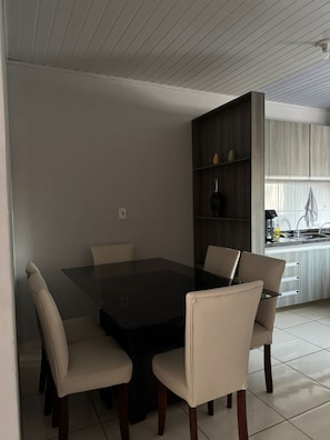 Ein stilvoller Essbereich mit einem Tisch mit Glasplatte und vier Stühlen, einem dunklen Holzregal und einer Küchenzeile mit Spüle und Schränken.
