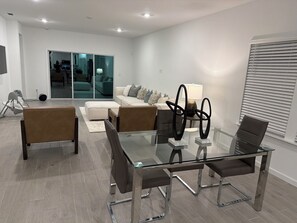 Un salon moderne avec une table à manger en verre, des fauteuils en cuir, un canapé et une grande fenêtre avec des stores.