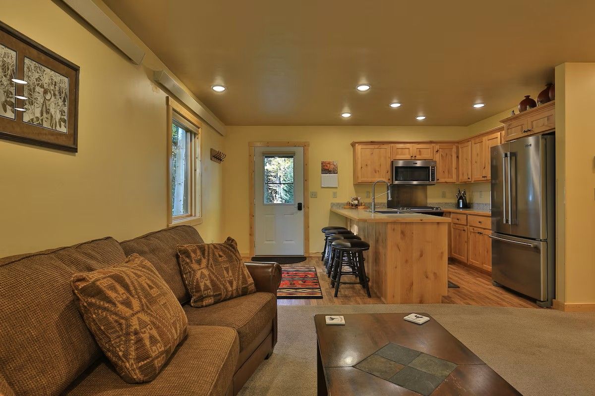 A cozy living room with a sofa and a coffee table, a kitchen with wooden cabinets and stainless steel appliances, and a door leading to a bright area.