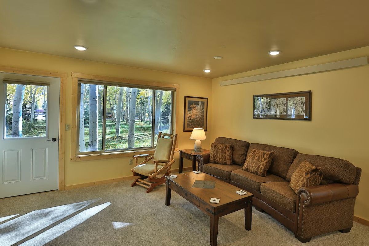 A living room with a sofa, armchair, coffee table, and a door leading to a view of trees.