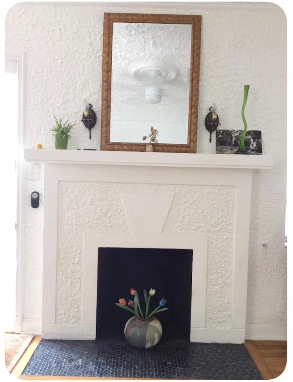 Una chimenea blanca con un espejo decorativo encima, un estante pequeño con varios objetos y un jarrón con flores en la base.