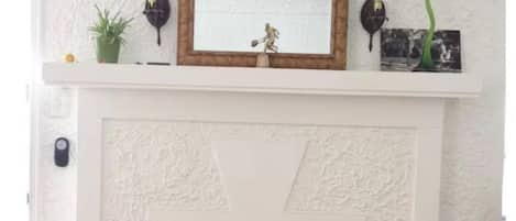 A white fireplace with a decorative mirror above it, a small shelf with various items, and a vase with flowers at the base.