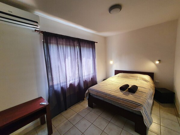 Un dormitorio pequeño y ordenado con una cama individual, una mesita de noche de madera, un escritorio y una ventana con cortinas.