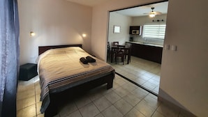 A bedroom with a bed, a bedside table, a chair, and a view into a kitchen area. - Marina Loft Studio Number 223 (Sardinal)