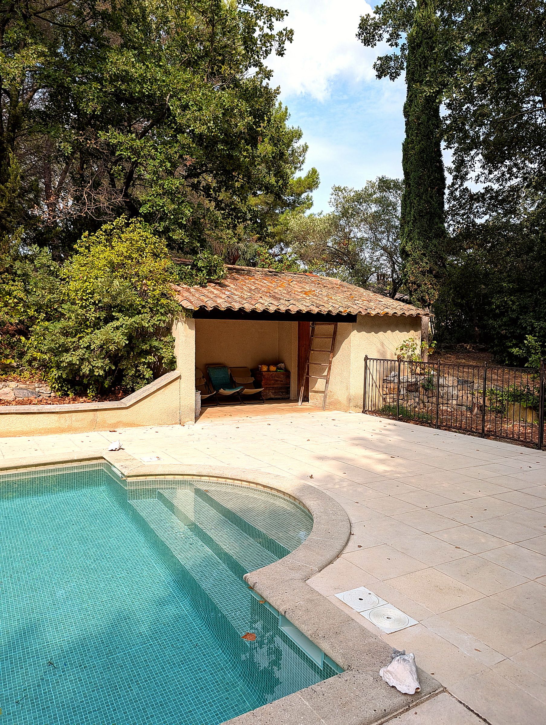 A swimming pool with a tiled deck, a covered seating area, and a fenced garden.