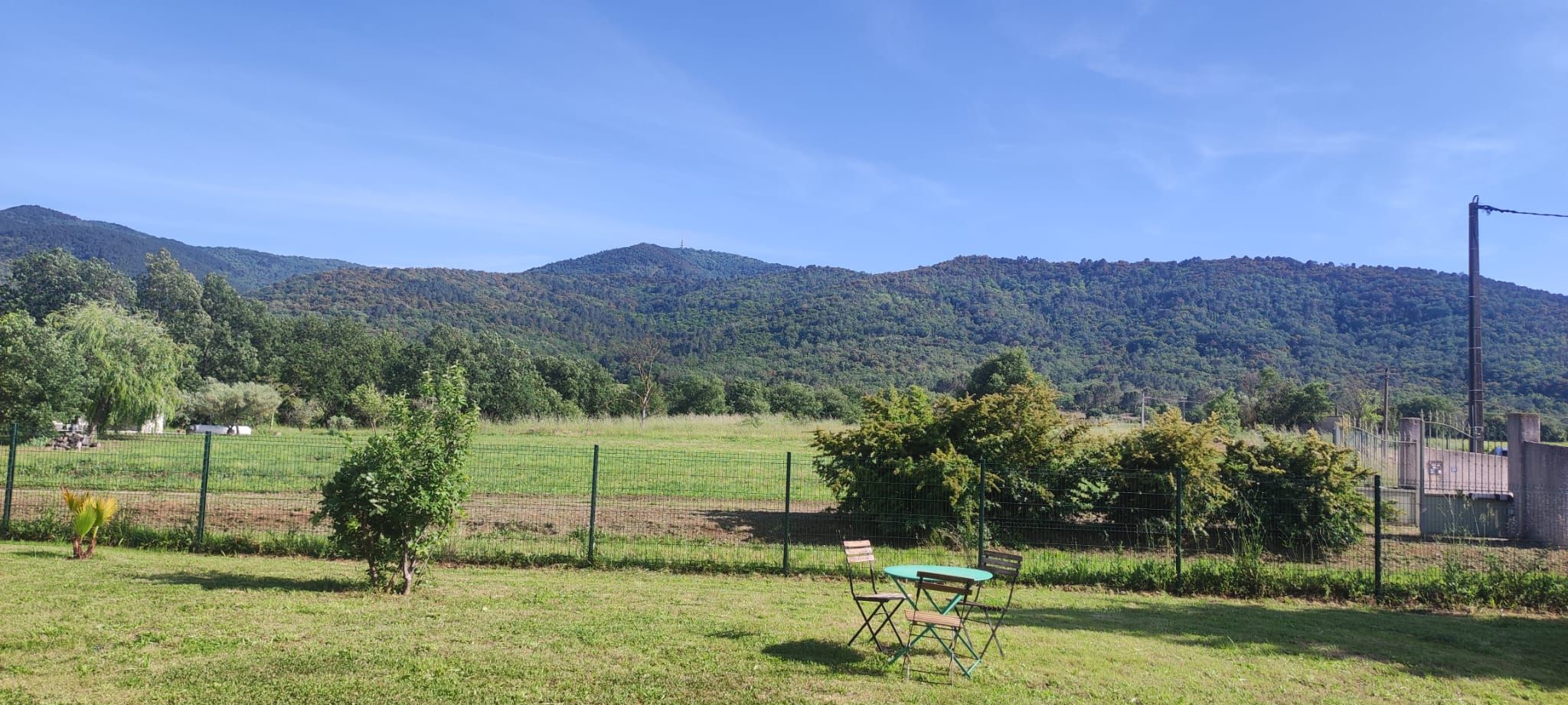 Un champ verdoyant avec une table et des chaises, entouré d’une clôture et d’arbres, avec des montagnes en arrière-plan.