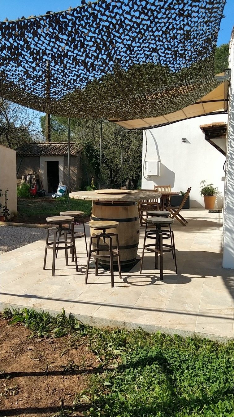 Een terras buiten met een ronde houten tafel, vier krukken en een barbecue.