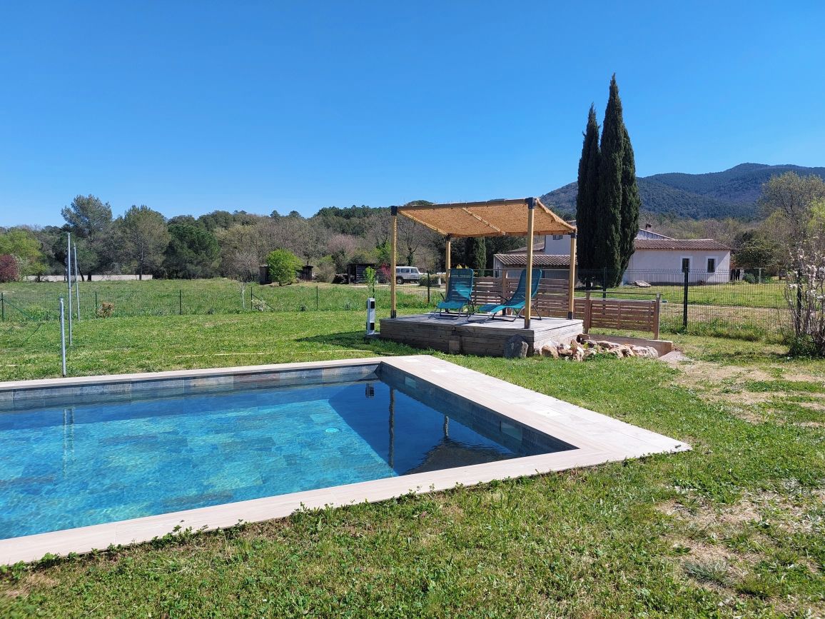 Une piscine dotée d’une terrasse en bois et de chaises longues, entourée d’une pelouse et d’une propriété clôturée.