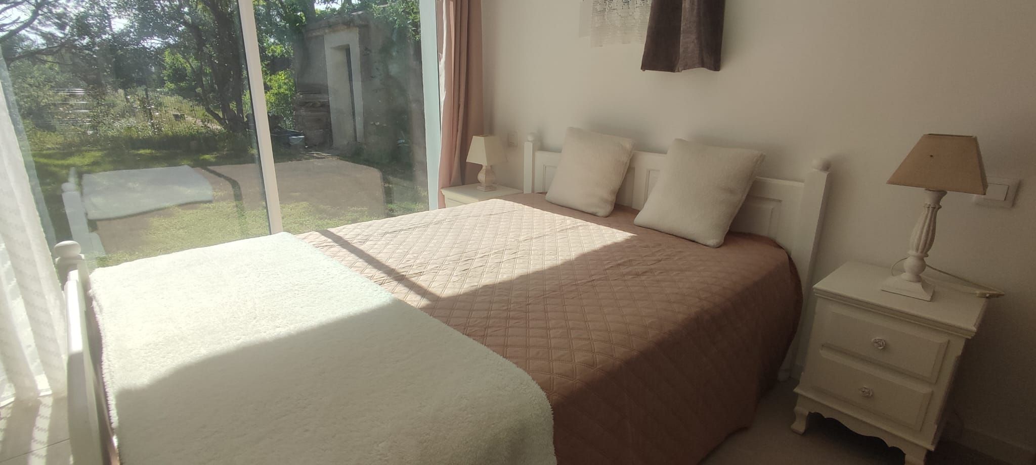 Een slaapkamer met een groot bed, een nachtkastje met een lamp, en uitzicht op de tuin door een glazen deur.