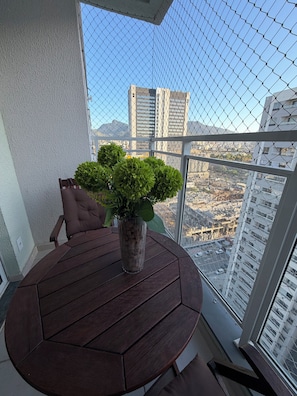 Ein runder Holztisch mit einem Blumentopf, ein brauner Stuhl und Blick auf eine Stadtlandschaft durch ein Maschendrahtgitter.