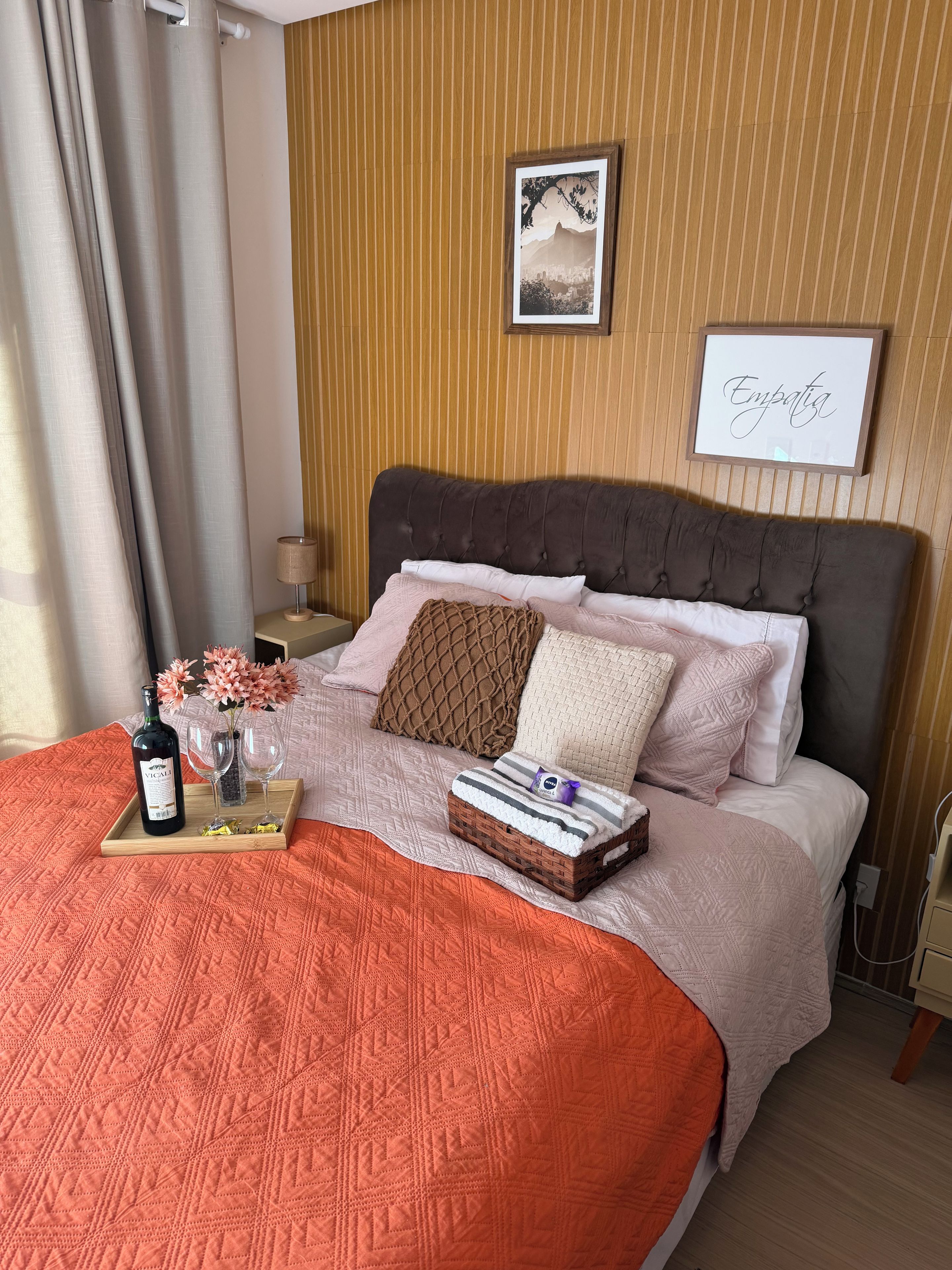 A neatly made bed with a brown headboard, a framed picture on the wall, and a tray with a bottle and glasses on the bed.