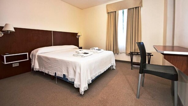 A hotel room with a bed, a desk, a chair, a lamp, and a window with curtains. - Hotel Playa (Comodoro Rivadavia)