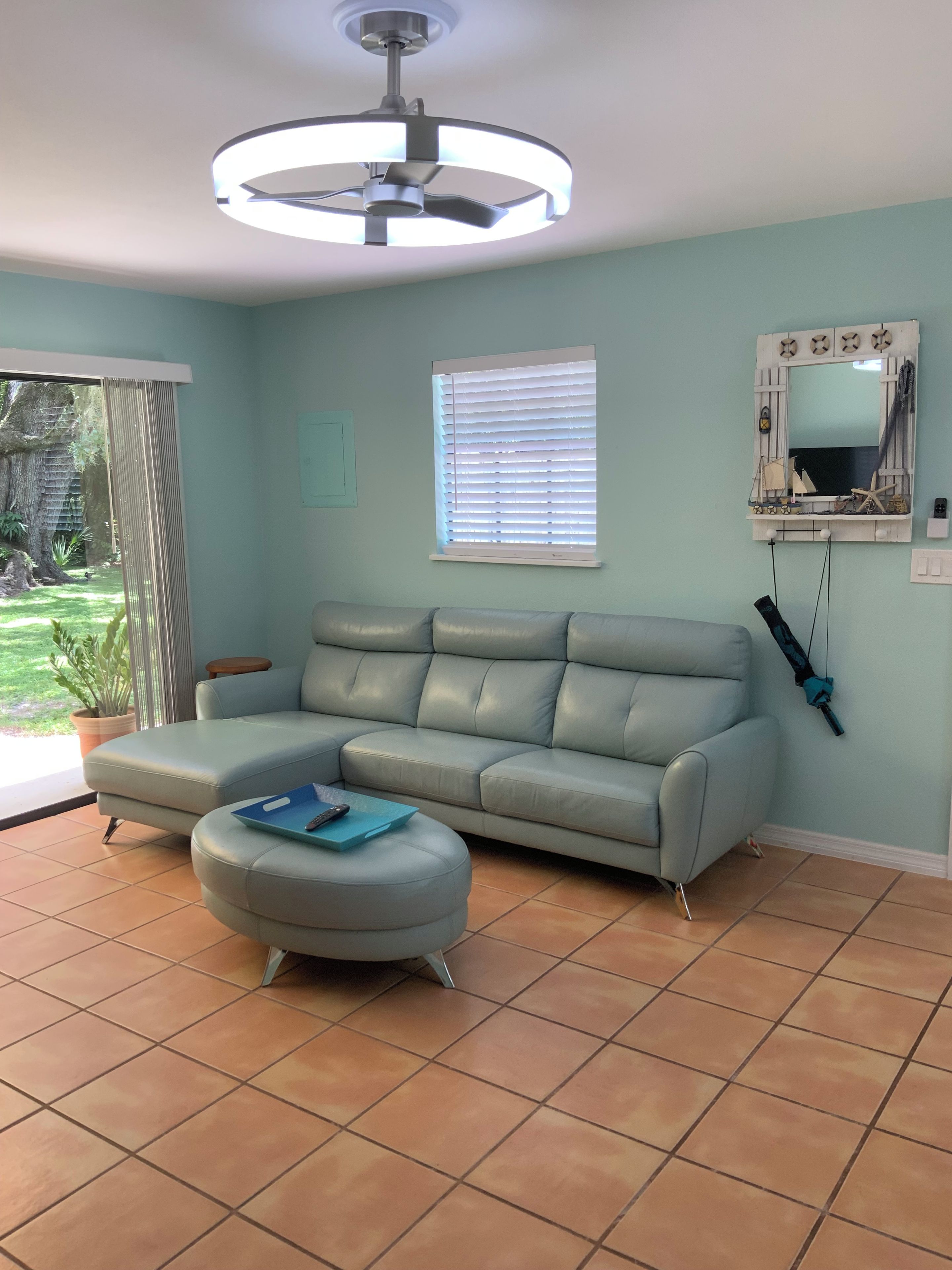 A living room with a grey sofa, a matching ottoman, a ceiling fan, a window with blinds, and a wall-mounted TV.