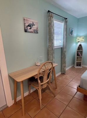 A room with a wooden table and chair, a window with curtains, a wall with a framed picture, and a shelf with books and a lamp.